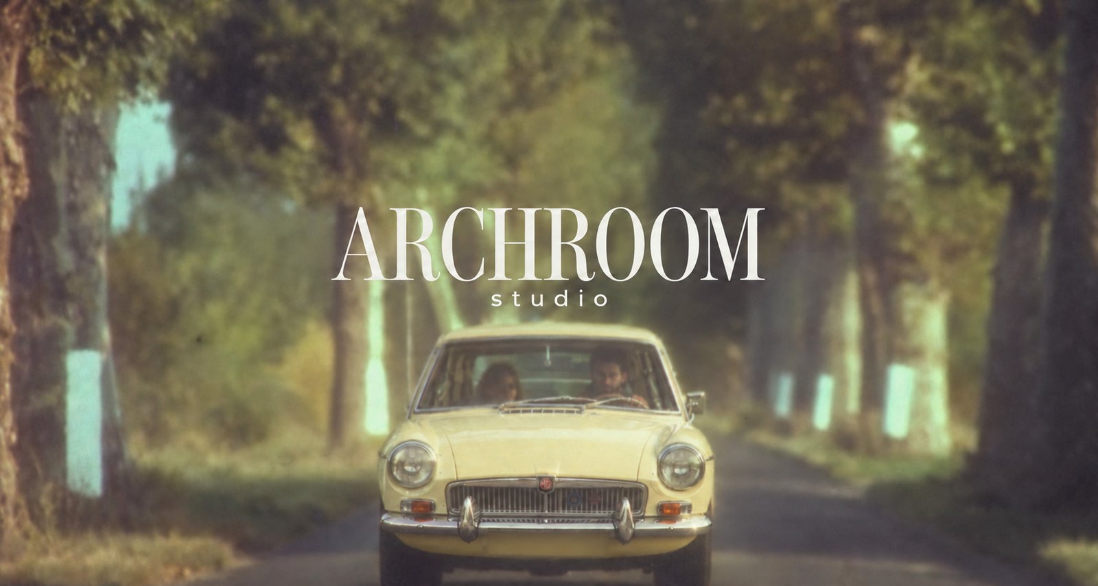 ARCHROOM studio above a pale yellow vintage car, with two people inside, on a tree-lined road.